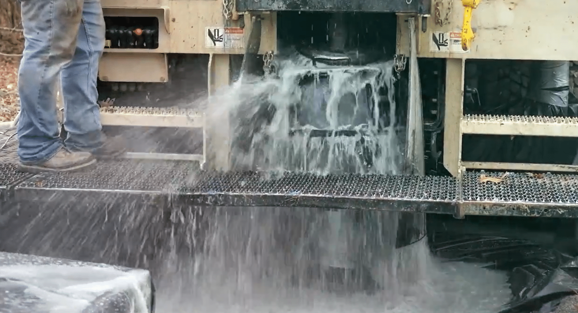 Water pours over metal grates from a machine in an industrial process, splashing down. A worker's legs in jeans and boots appear on left.