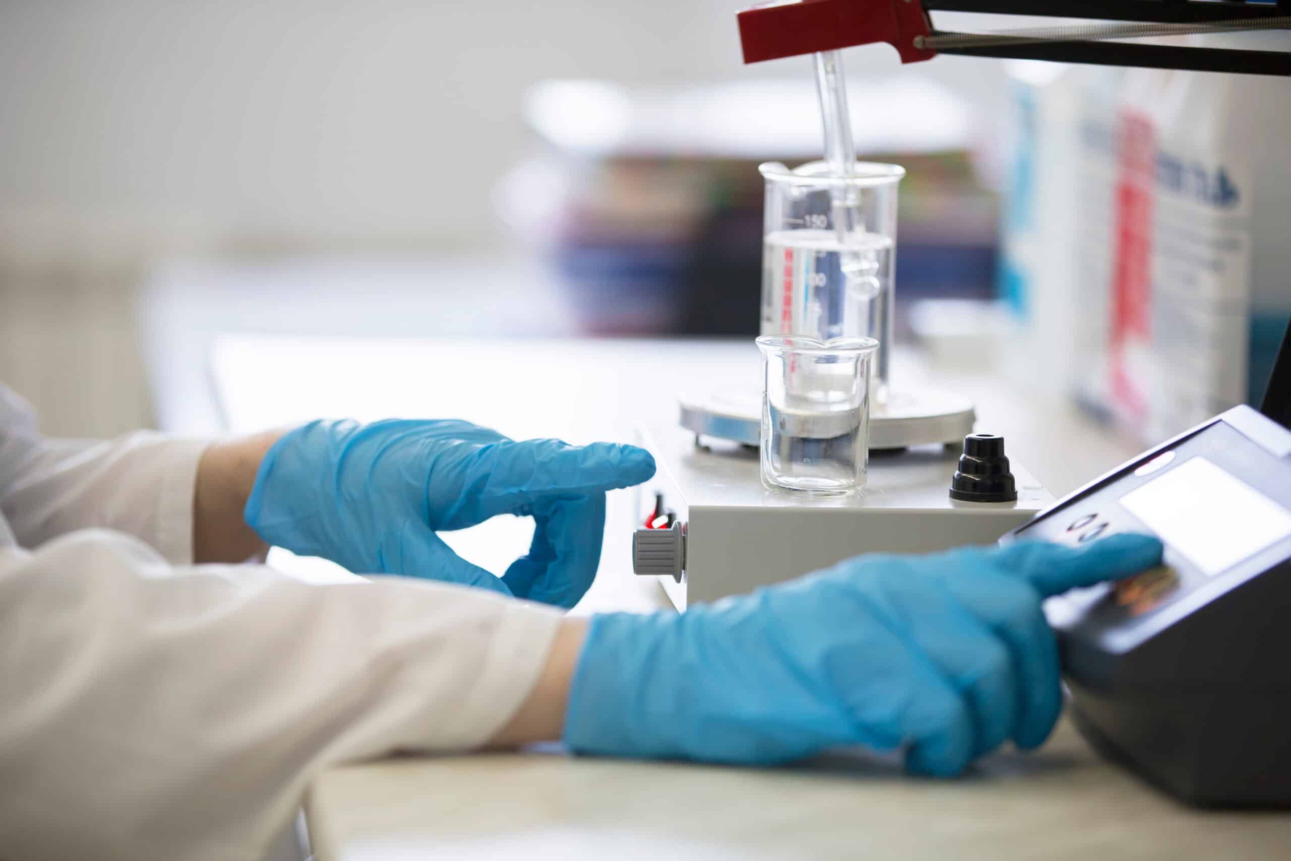 A person in blue gloves uses lab equipment, with beakers and glassware on a magnetic stirrer in a brightly lit laboratory.