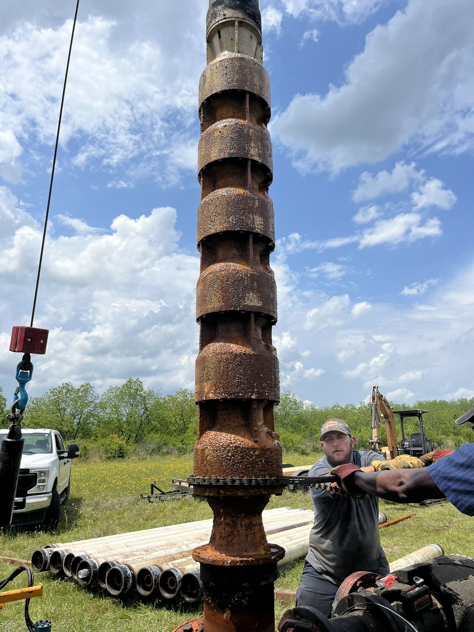 Two workers beside a large rusty cylindrical metal pump being lifted by machinery on a grassy field, with equipment and truck behind.