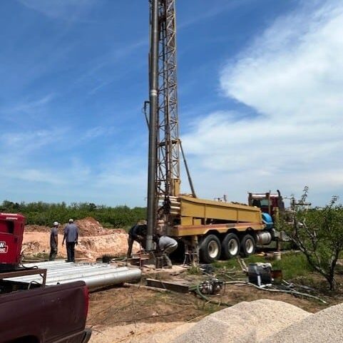 A tall water well drilling rig with workers on a dirt site, pipes and equipment nearby, and a red truck in the foreground under a blue sky.