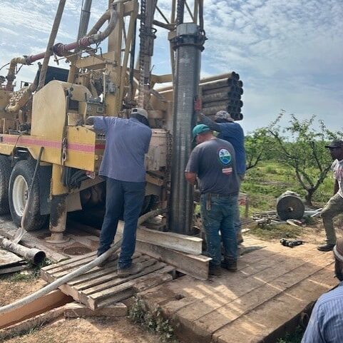 Workers operate a large drilling rig outdoors in sunlight, handling pipes and equipment on a wooden platform amid dirt and tools.