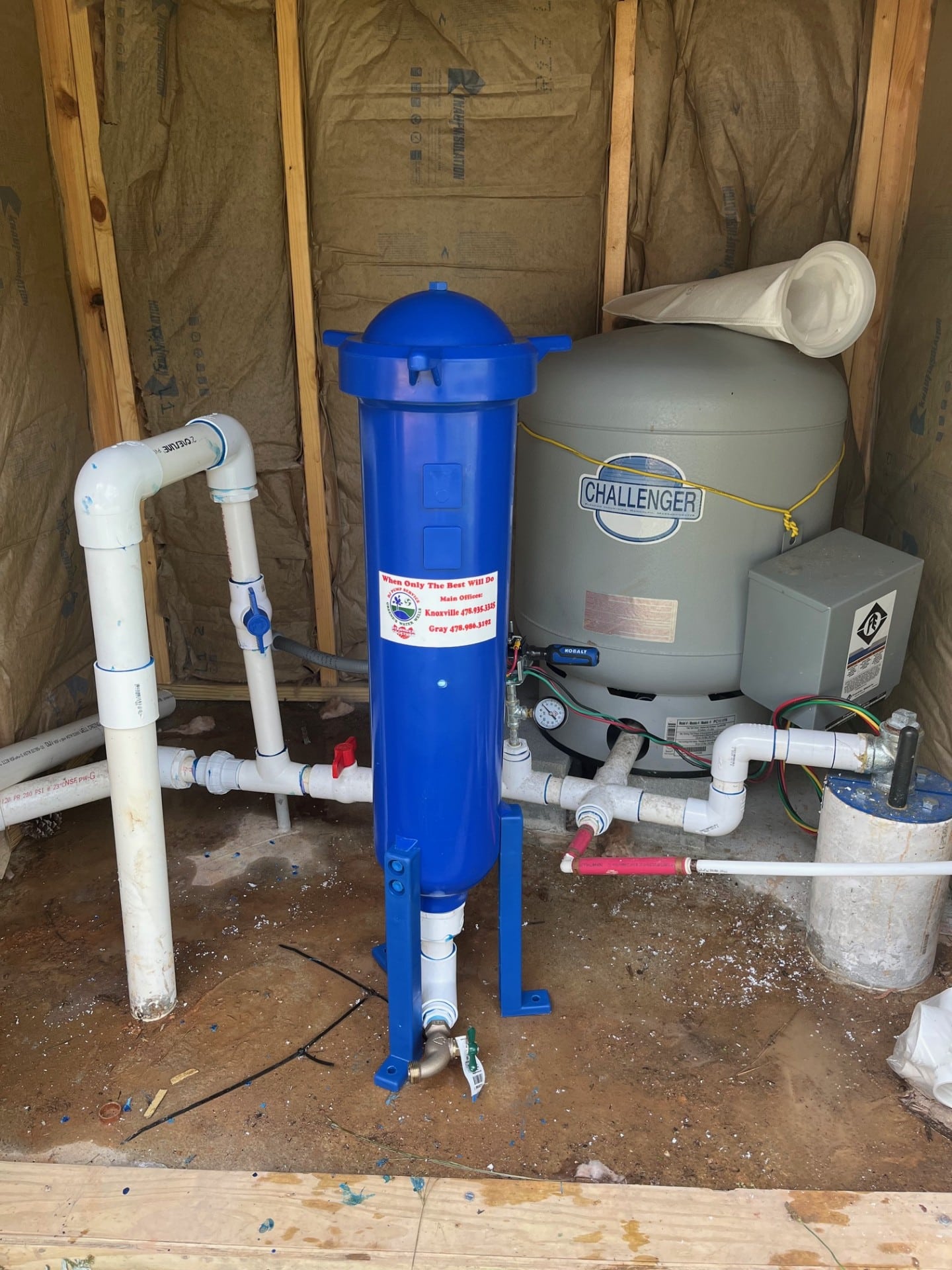 A blue water filter system on a concrete floor in an unfinished space with exposed insulation, near a gray Challenger water tank and PVC pipes.