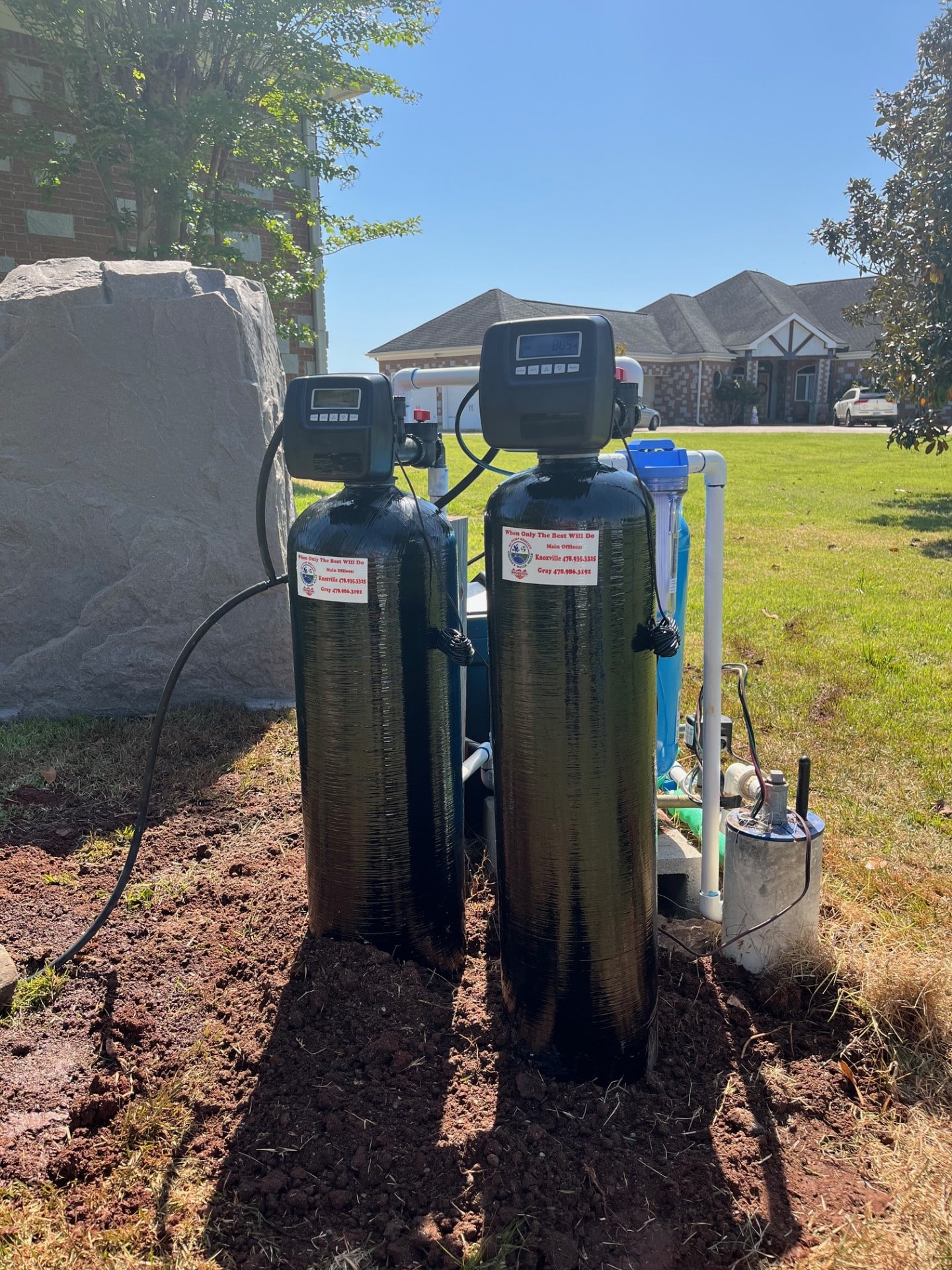Two black water softener tanks with digital meters outdoors on dirt, near pipes and blue filter; houses and rock under clear sky.