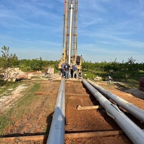 Three workers by a tall drilling rig in a grassy field, next to large metal pipes under a clear blue sky.