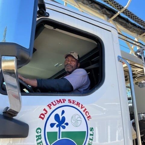 A man wearing a cap sits in the driver's seat of a truck labeled DJ Pump Service Greene's Water Wells. The white truck is parked outdoors on a sunny day.