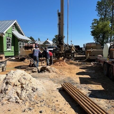 Two workers drill at a construction site by a green house. Metal pipes, machinery, and trees are visible under a clear blue sky.