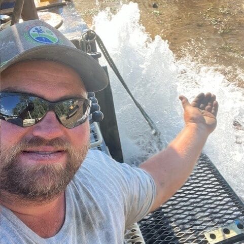 A bearded man in sunglasses, a cap, and gray shirt smiles on metal grating beside a powerful stream of water from outdoor machinery.