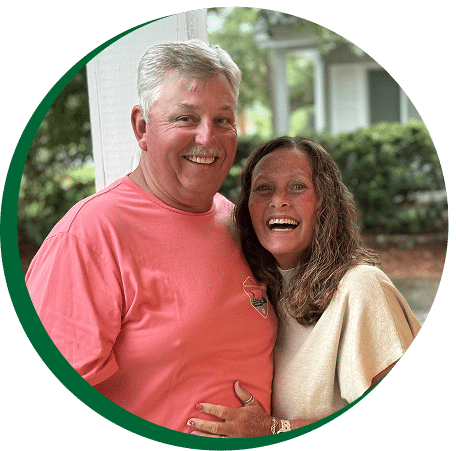 A smiling man in a pink T-shirt stands beside a woman with curly brown hair and a beige top outdoors, greenery and house behind them.