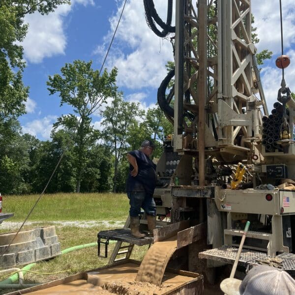 A worker on a drilling rig platform watches muddy water flow down a chute; another person is nearby. Green trees and blue sky behind.