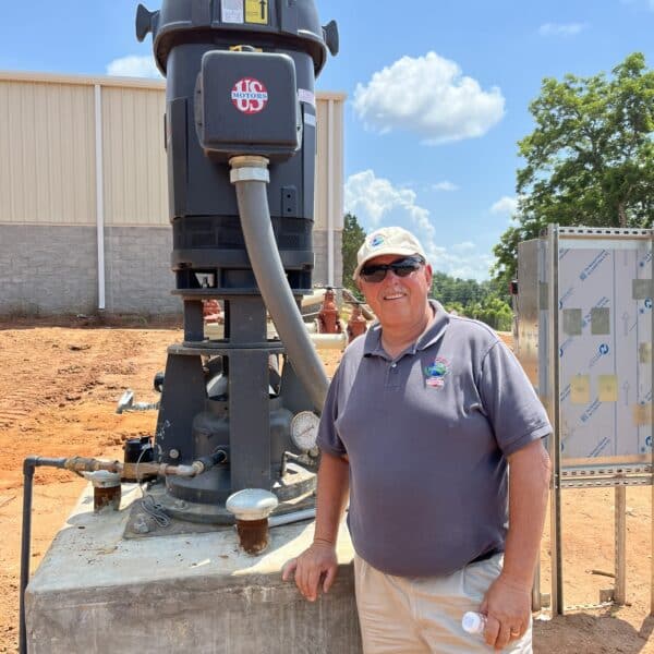 A man in sunglasses and a gray polo shirt stands outdoors by industrial equipment; behind him are a building, sky, and trees.