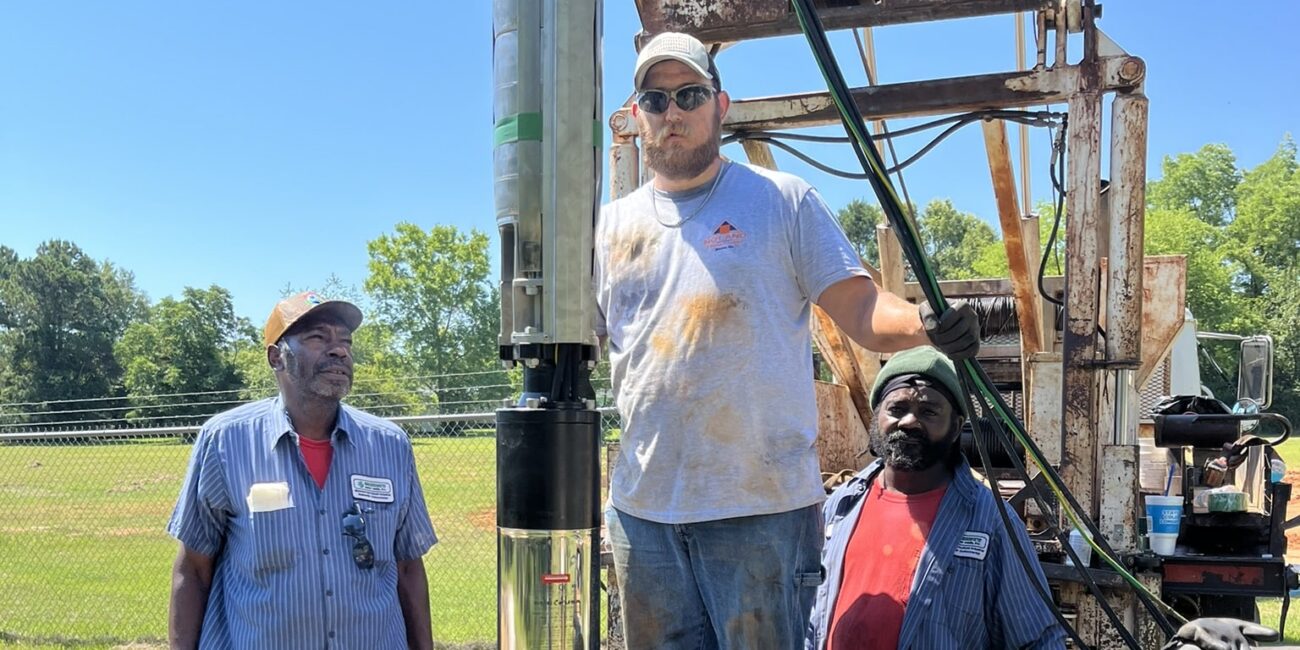 Three workers at a well site with machinery; one holds a large pipe on the well edge while others watch on a sunny outdoor day.