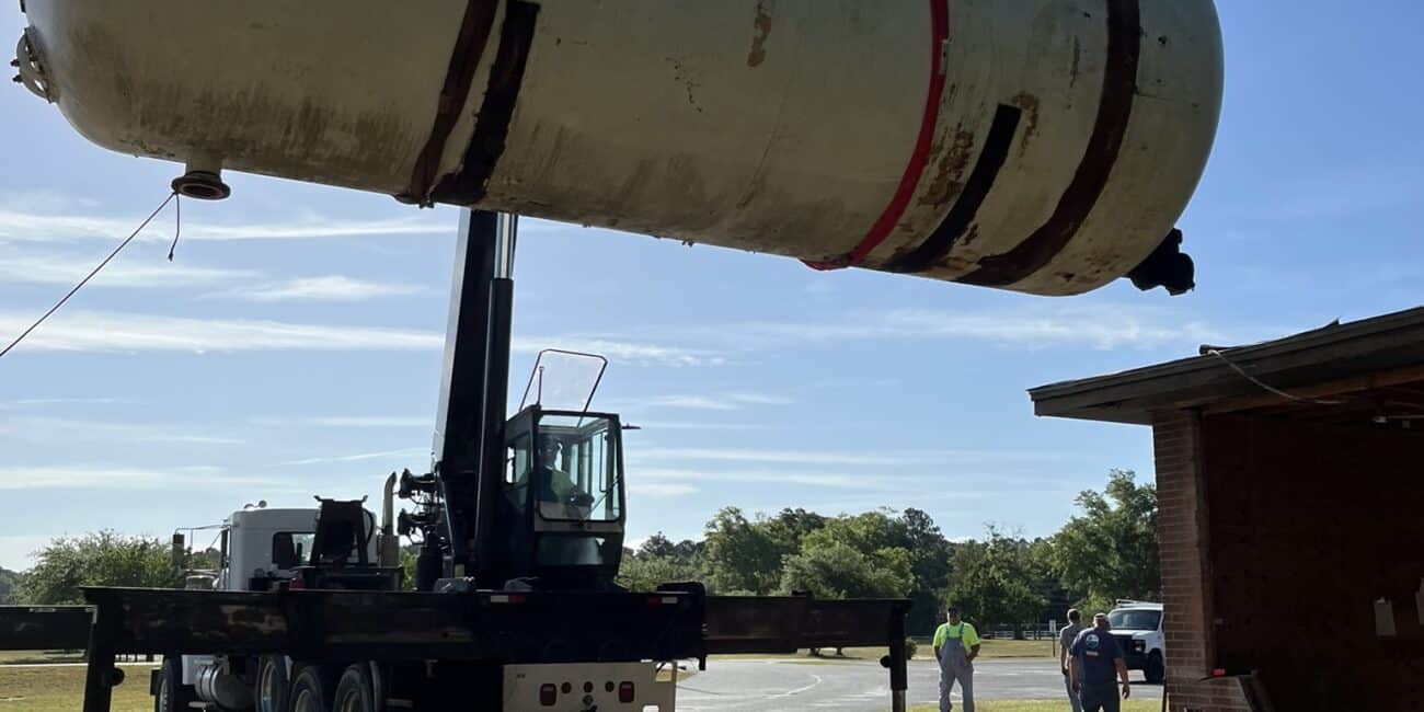 A large crane lifts a cylindrical storage tank with red straps near a brick building as people stand on a grassy area under blue sky.