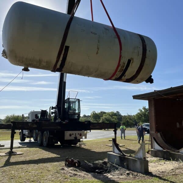 A large crane lifts a cylindrical storage tank with red straps near a brick building as people stand on a grassy area under blue sky.