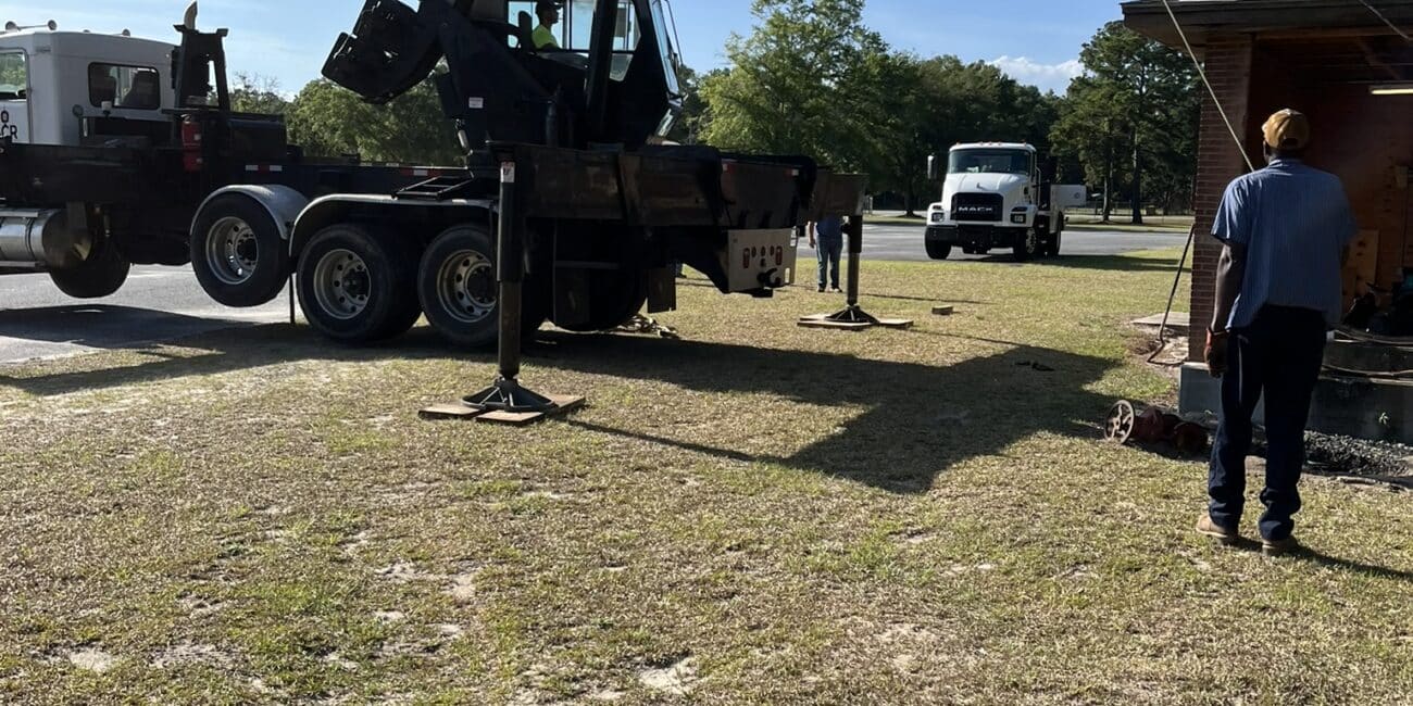 A crane labeled Batista Crane lifts a large cylindrical tank near a small building, while several workers stand nearby on grassy ground.