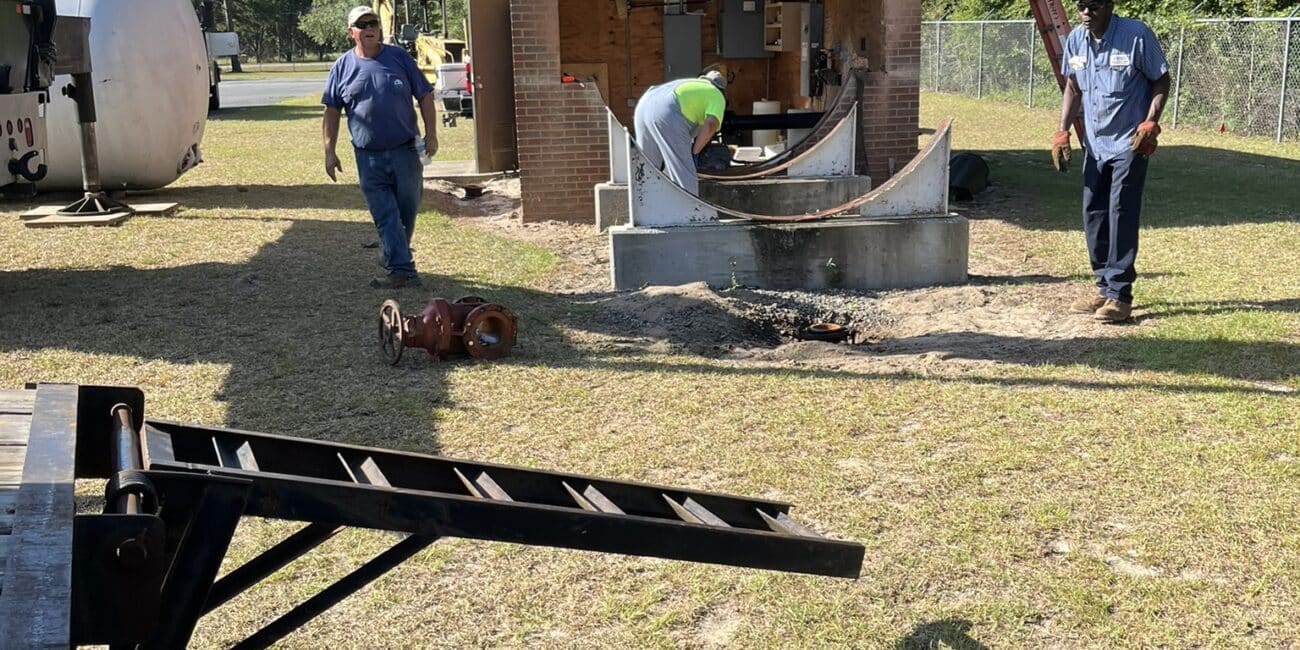 Three people work by a brick building with construction materials and gear on the grass. Trees, fence, and blue sky behind.