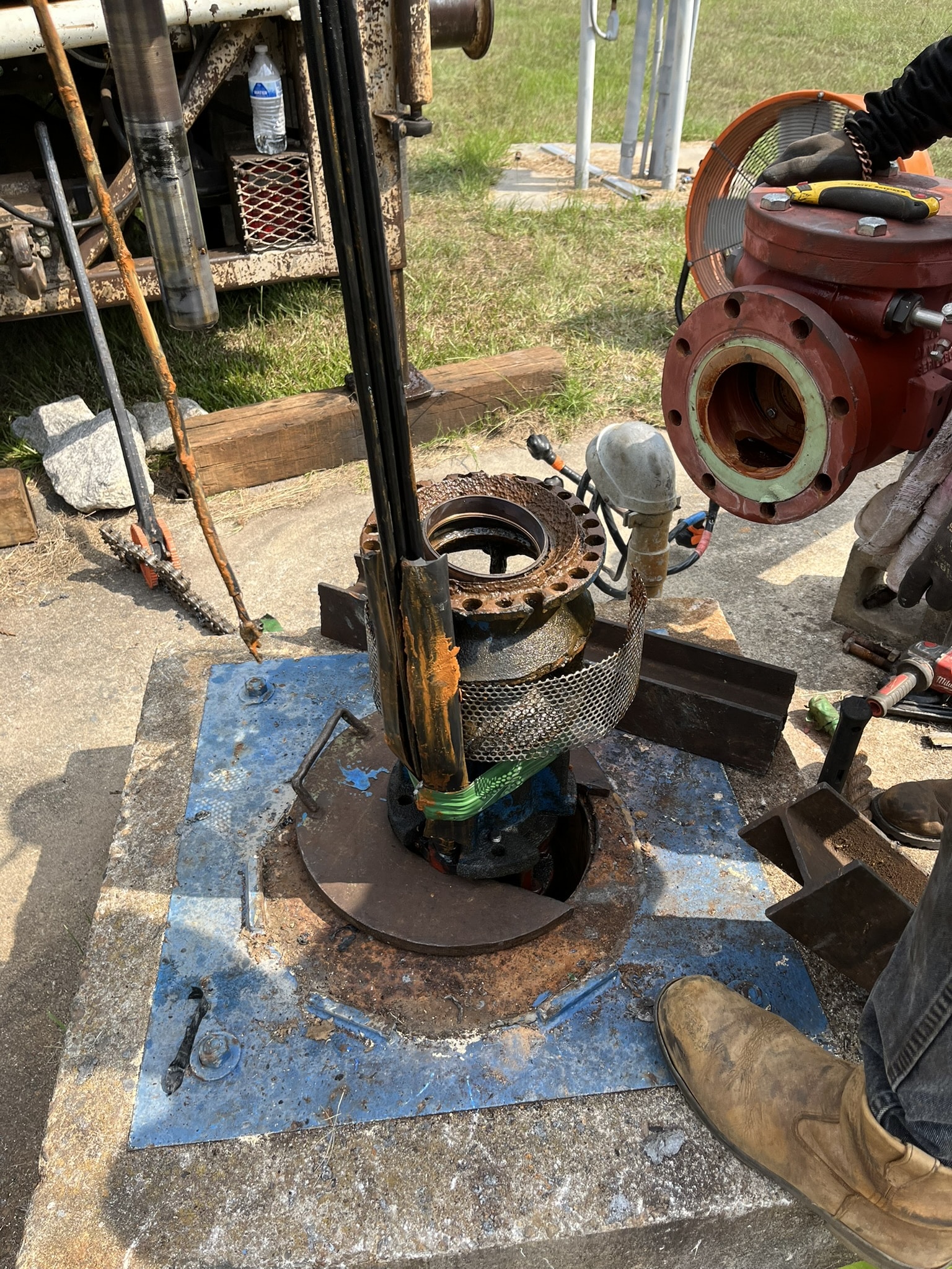 Close-up of outdoor industrial equipment being serviced; pipes, valves, fittings, a boot on a rusty blue platform with tools scattered.