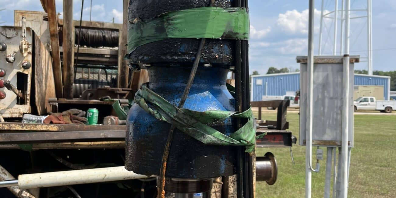 Close-up of blue and black machinery with green tape at an industrial site; water tower and buildings in the background, cloudy sky.