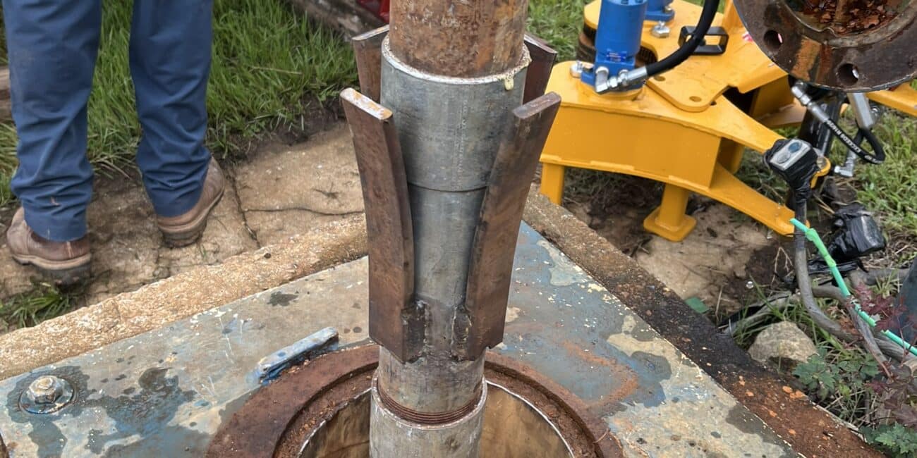 Close-up of a metal pipe being lowered into a well on a weathered plate, with a person and mechanical equipment visible in the background.