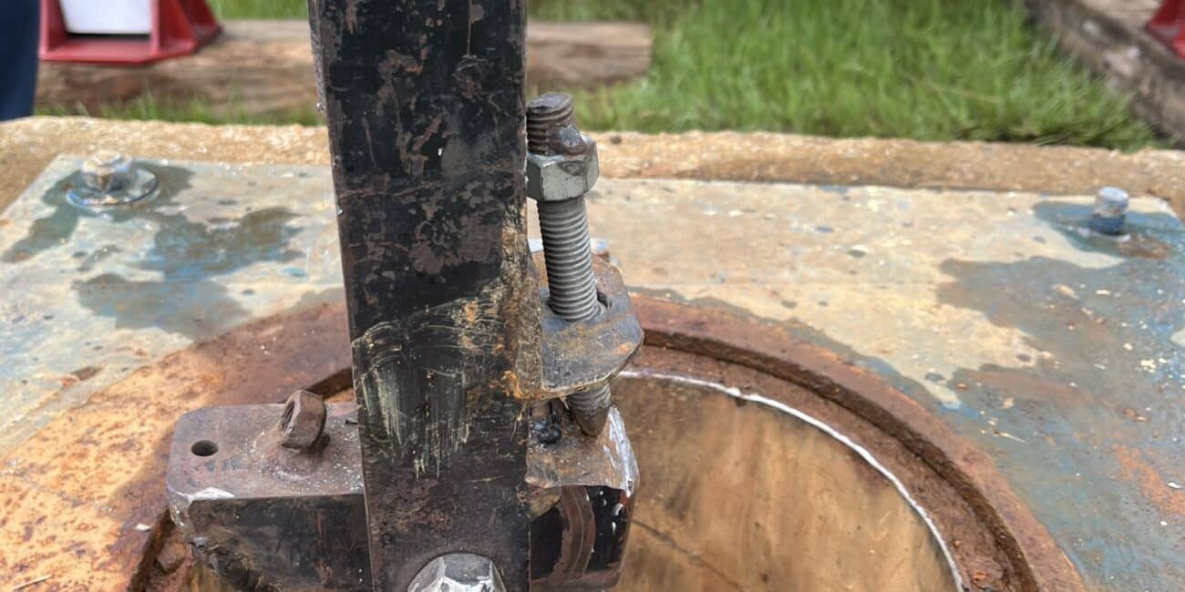 Close-up of a rusted metal mechanism on the rim of a round opening in a thick plate, blue truck and grass blurred in background.