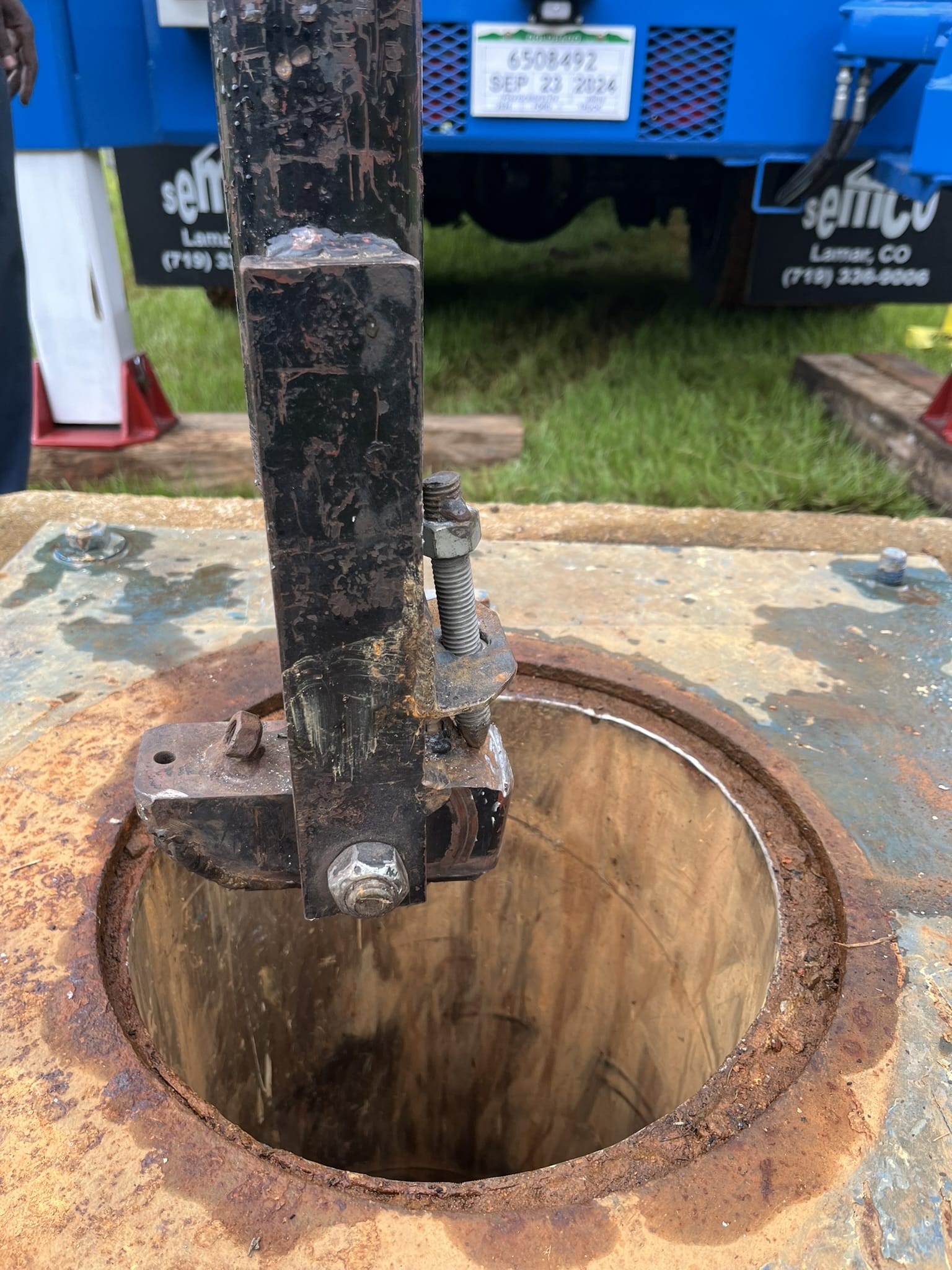 Close-up of a rusted metal mechanism on the rim of a round opening in a thick plate, blue truck and grass blurred in background.