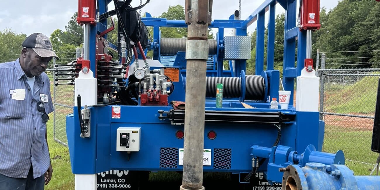 A worker stands by a large blue drilling rig over an open well on grass, with equipment, cables, a fence and trees in the background.