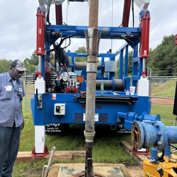 A worker stands by a large blue drilling rig over an open well on grass, with equipment, cables, a fence and trees in the background.