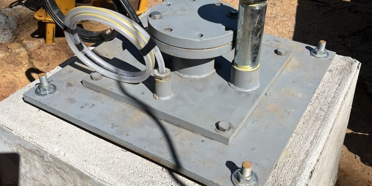 A metal hydraulic jack and hoses sit on a concrete block at a construction site, sand and equipment in the sunlit background.
