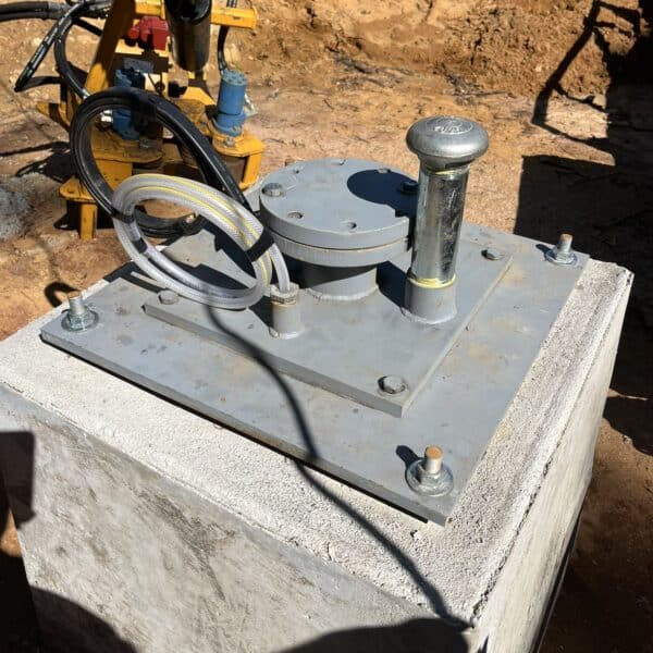 A metal hydraulic jack and hoses sit on a concrete block at a construction site, sand and equipment in the sunlit background.