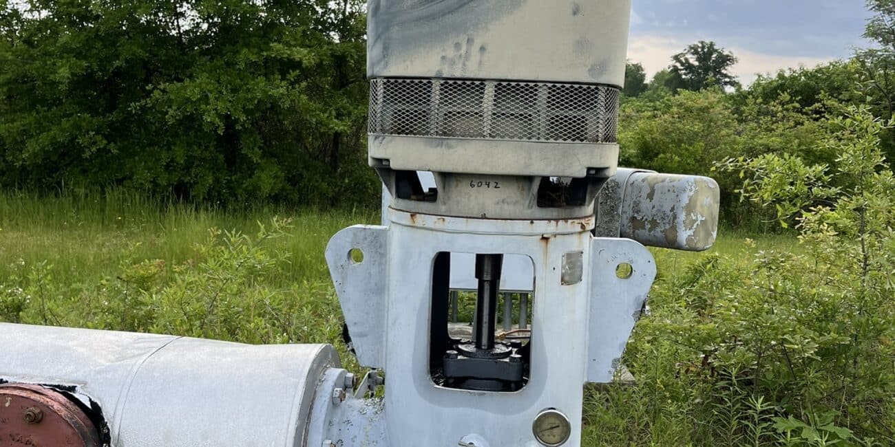 A large, weathered industrial pump with a cylindrical motor sits on a concrete platform outdoors amid grass and trees under clouds.