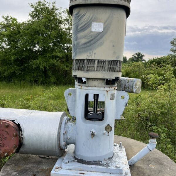 A large, weathered industrial pump with a cylindrical motor sits on a concrete platform outdoors amid grass and trees under clouds.