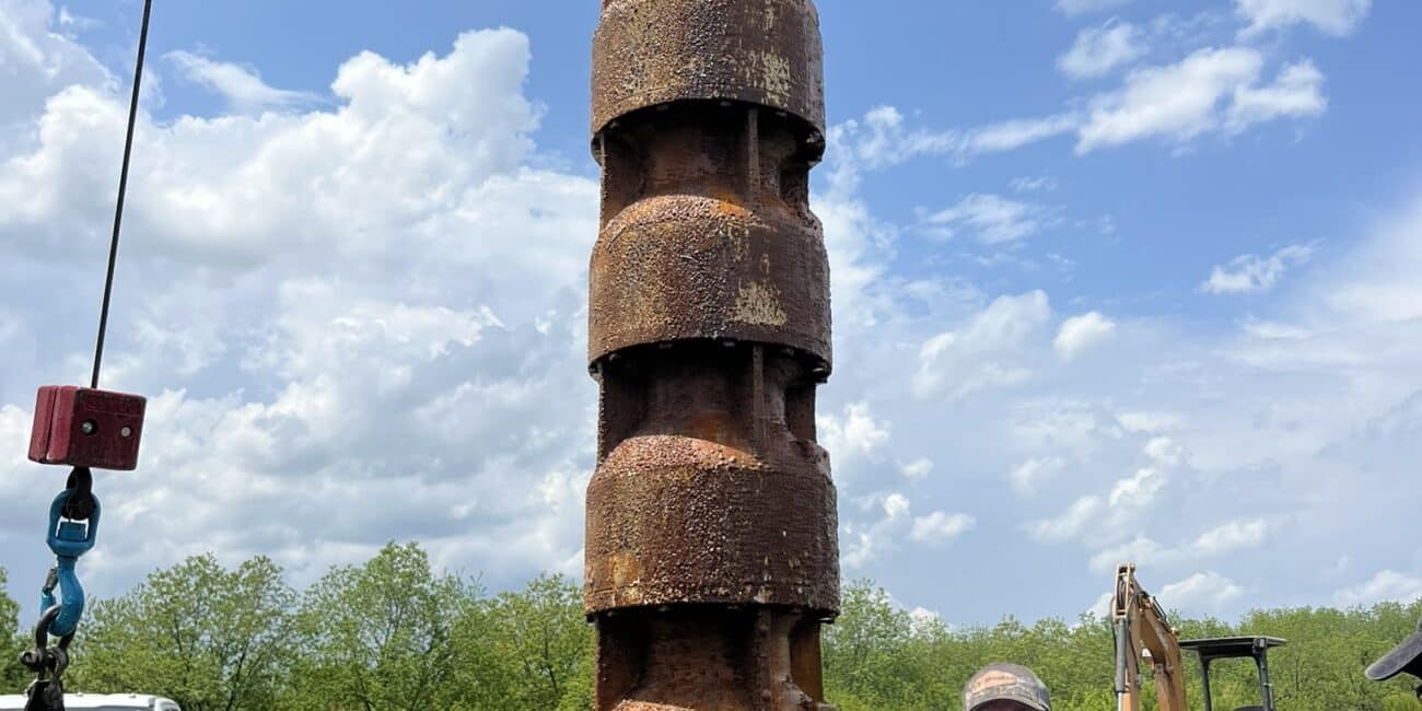 A rusty industrial pump is lifted by a crane outside as two workers guide it, with pipes and machinery on grass under a cloudy sky.