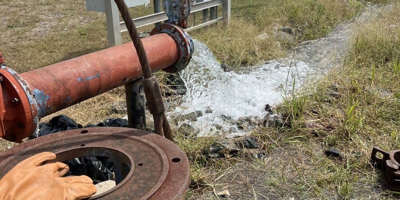 Water pours from a big red pipe onto grass near utility boxes and trucks, with rusted gear and gloves hinting at outdoor repairs.