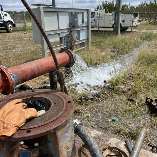 Water pours from a big red pipe onto grass near utility boxes and trucks, with rusted gear and gloves hinting at outdoor repairs.