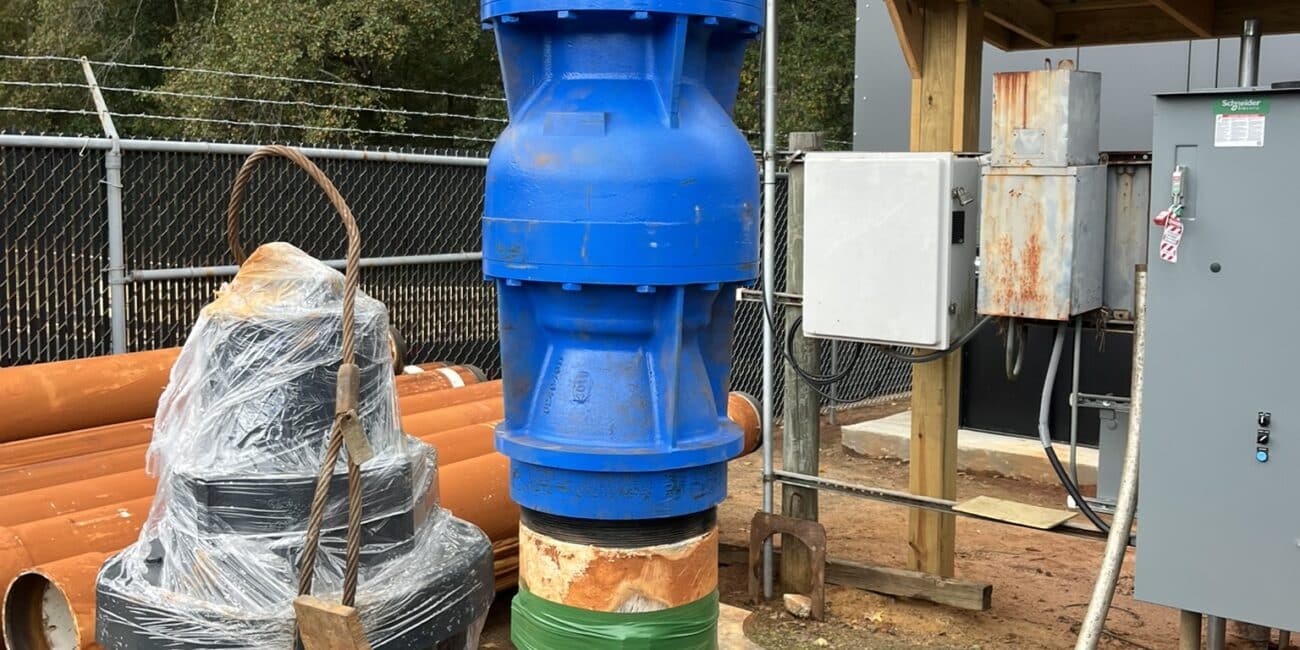 A large blue industrial pipe assembly stands vertically outdoors, supported by wooden braces, with control panels, orange pipes, and trees nearby.