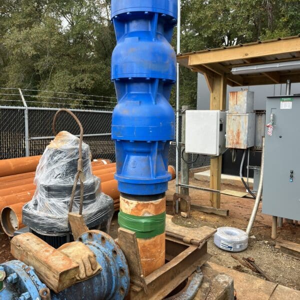 A large blue industrial pipe assembly stands vertically outdoors, supported by wooden braces, with control panels, orange pipes, and trees nearby.