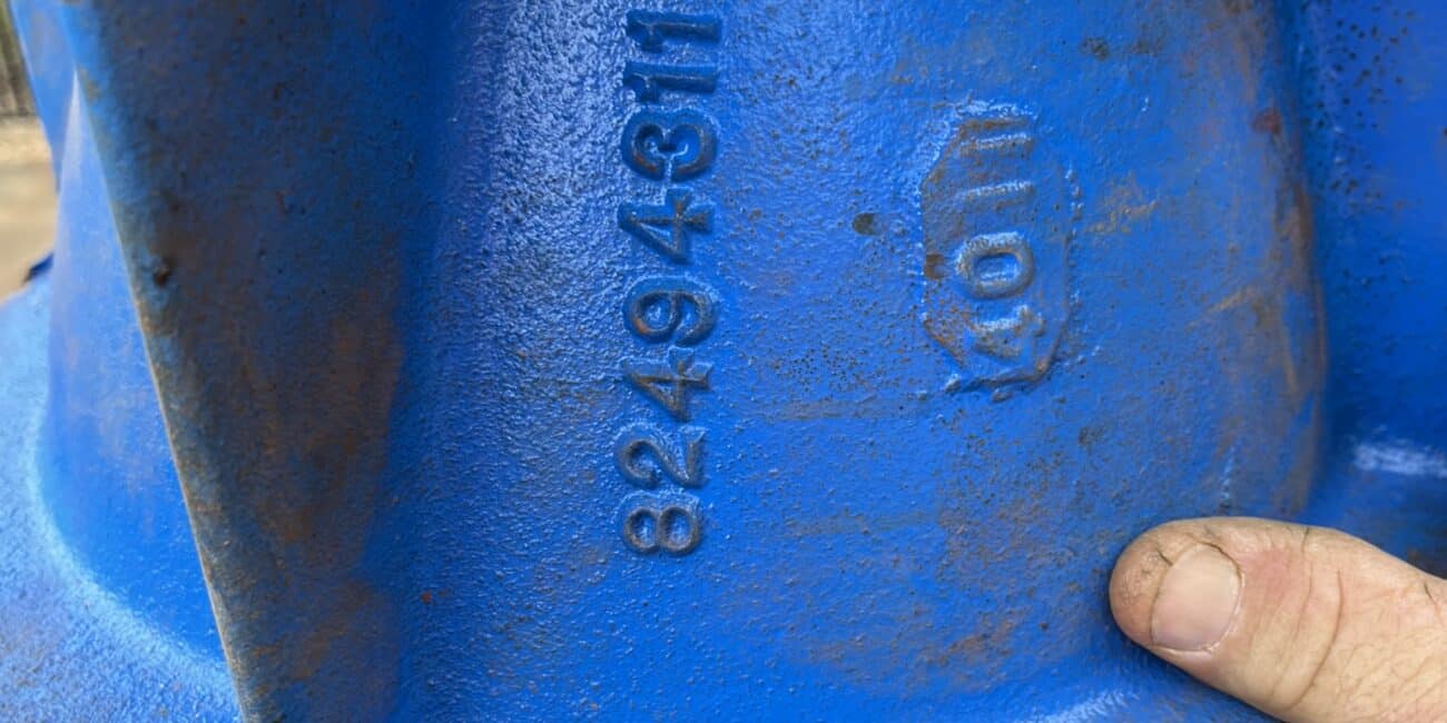 A close-up of a blue industrial metal part with numbers 8249431 and 10M embossed. A thumb holds the part, which shows rust and dirt.