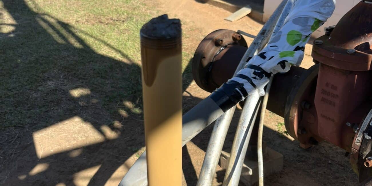 Close-up of a cylindrical metal rod coated with brown substance, attached to outdoor industrial machinery; utility vehicles and pipes behind.