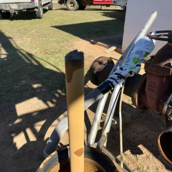 Close-up of a cylindrical metal rod coated with brown substance, attached to outdoor industrial machinery; utility vehicles and pipes behind.