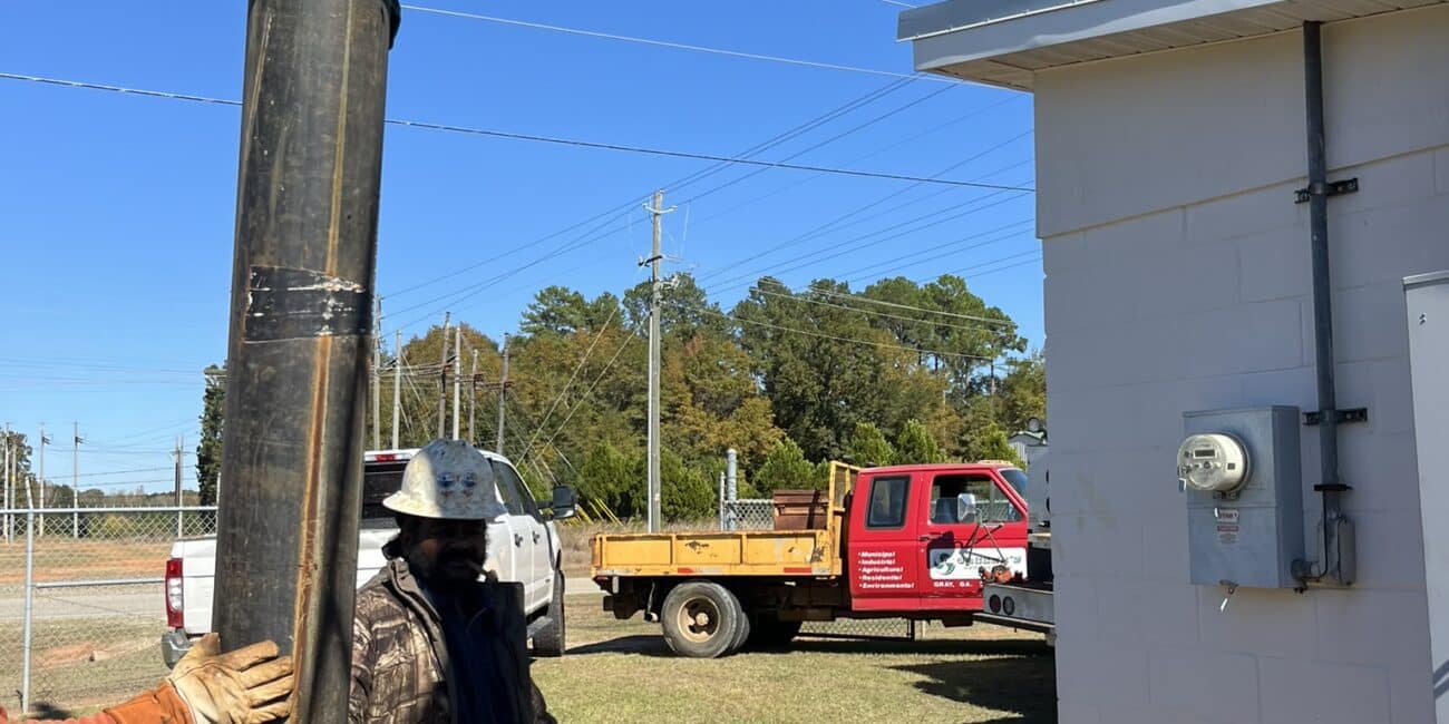 Two workers use a hoist to move a large blue industrial pipe near a white building, with vehicles and trees under a clear sky.