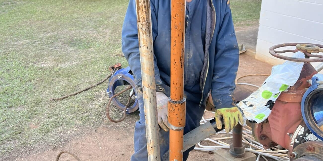 A worker in safety gear stands behind large orange and silver pipes from a round metal opening, doing maintenance near utility vehicles.