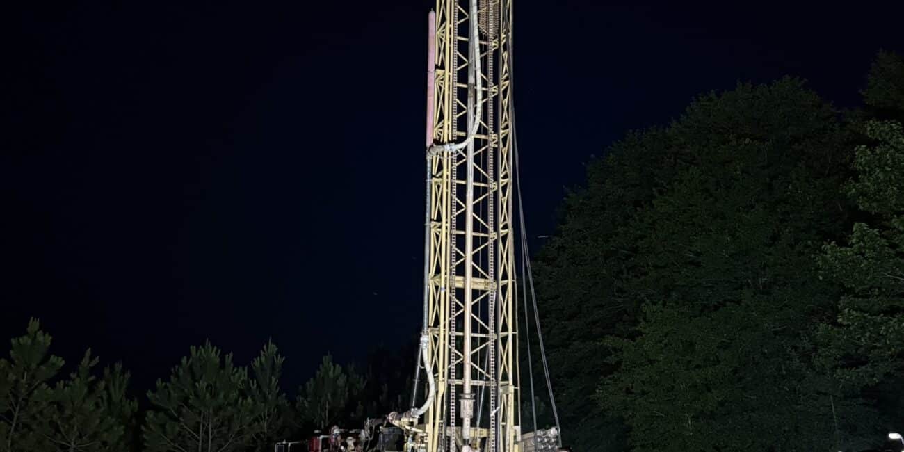 Tall illuminated drilling rig on reddish soil at night, yellow frame lit up amid equipment and trees against a dark forest sky.