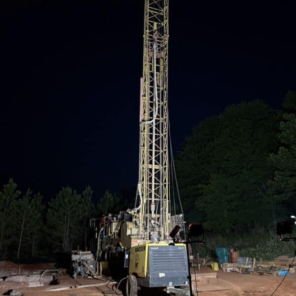 Tall illuminated drilling rig on reddish soil at night, yellow frame lit up amid equipment and trees against a dark forest sky.