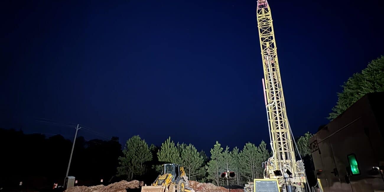 A night construction site lit by bright lights features a large yellow drilling rig, bulldozer, trees in back, and moon overhead.