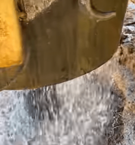 Close-up of an excavator bucket pouring water onto rocky ground, creating a splash as droplets scatter below.