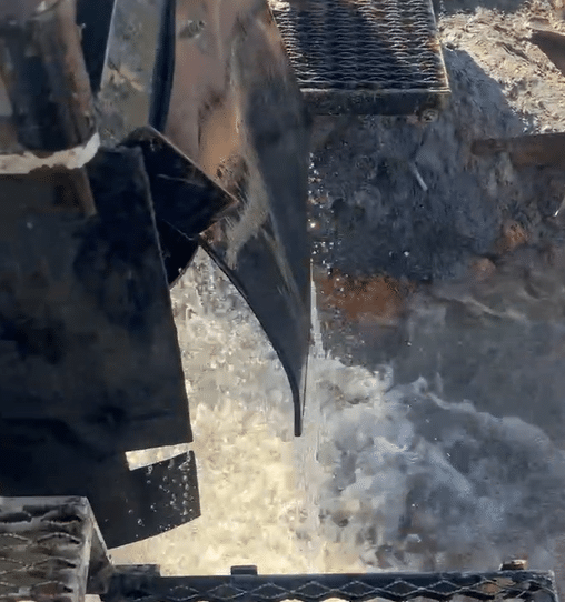 Water pours forcefully from a large metal chute onto rocks and gravel below, splashing. Grated platforms stand above the wet ground.