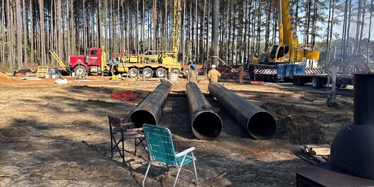 Construction site in a forest clearing: two cranes lifting pipes, red truck, metal pipes, blue chair, stove. Surrounded by pine trees.