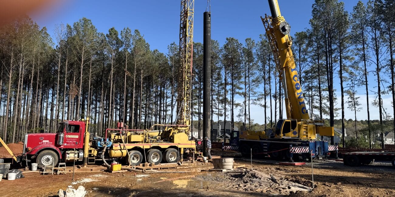 Construction site with yellow and red cranes lifting materials, pine trees behind. Clear blue sky. Finger partly covers top left corner.