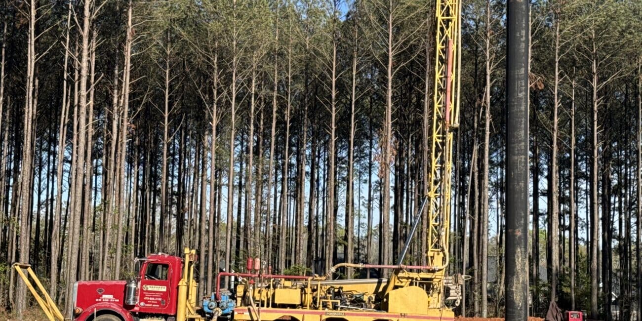 A large yellow drilling rig on a red truck operates at a muddy site with pine trees, construction materials, and equipment nearby.