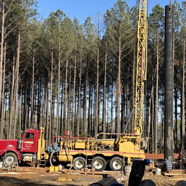 A large yellow drilling rig on a red truck operates at a muddy site with pine trees, construction materials, and equipment nearby.